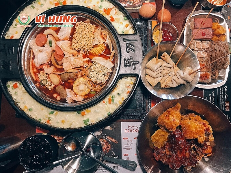 Nồi lẩu Hàn Quốc kiểu Dooka với vùng cháo trắng vòng ngoài và nước lẩu cay đỏ ở giữa, xung quanh bày sẵn bánh gạo, chả cá, miến và đồ chiên trên bàn tự phục vụ.