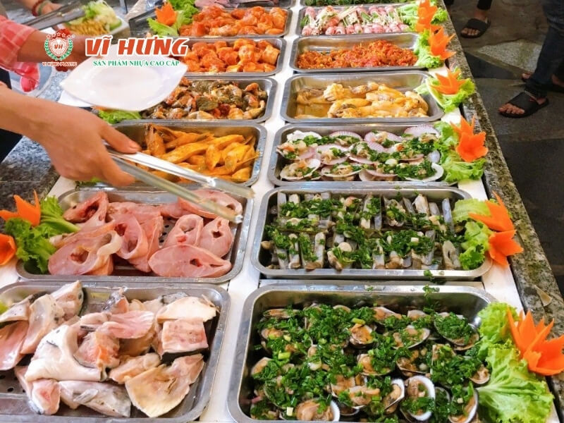 Quầy buffet tự chọn với nhiều khay inox đựng hải sản và món ăn chế biến sẵn, thực khách đang dùng kẹp gắp đồ ăn vào đĩa