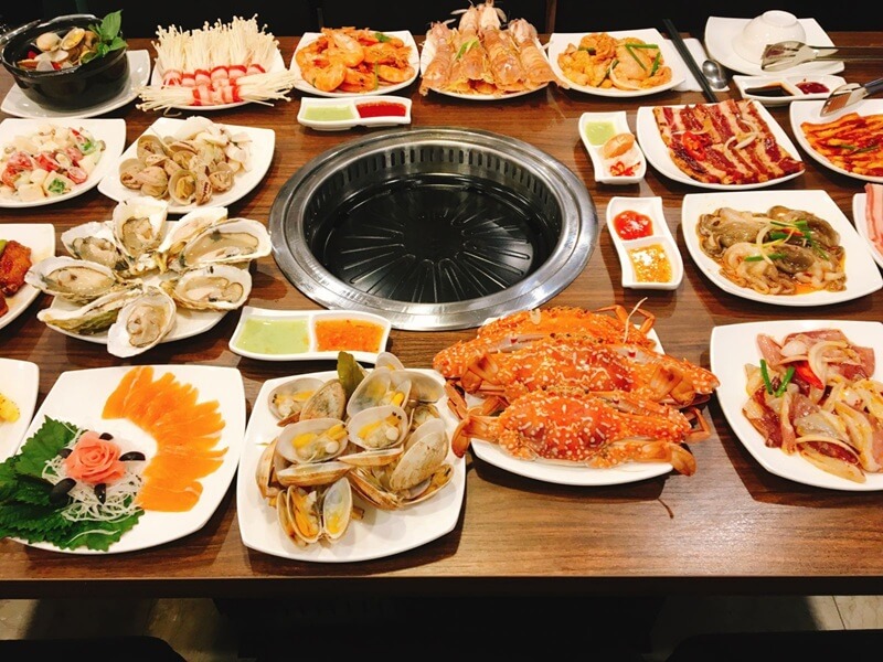 Bàn buffet hải sản đa dạng với sashimi cá hồi, hàu sống, nghêu, cua, tôm và bếp nướng than ở giữa