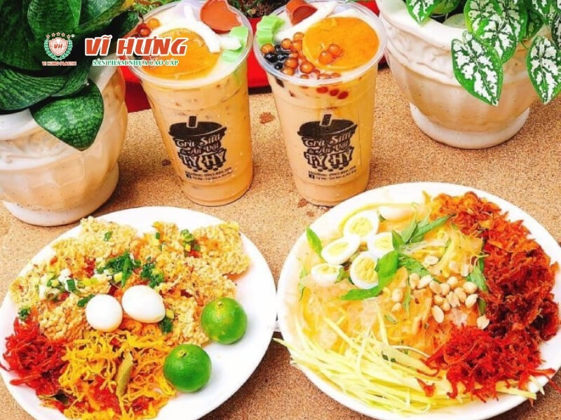 Ba ly trà sữa và hai đĩa đồ ăn vặt được sắp xếp trên nền cát, gồm trà sữa với topping colorful, và hai đĩa salad chua cay được trang trí bằng rau sống, trứng cút, và các loại rau thơm khác