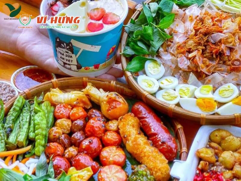 Một ly trà sữa màu xanh lam và nhiều đĩa đồ ăn vặt gồm các loại đậu edamame luộc, trứng cút nước mắm, chả cá, xúc xích nướng, rau sống như dưa chuột và bắp cải, sắp xếp trên mặt bàn