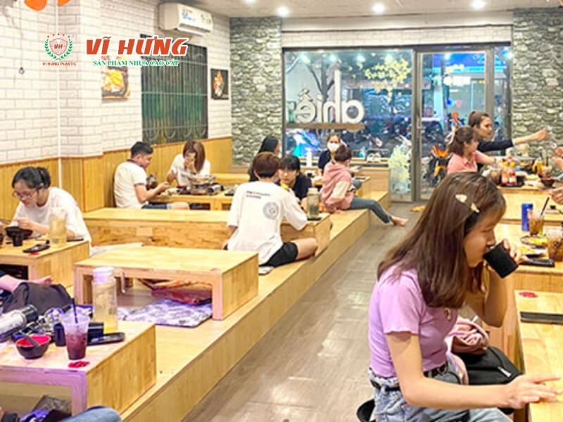 Quán trà sữa kết hợp đồ ăn vặt với không gian ngồi rộng rãi, bàn ghế gỗ sắp xếp gọn, khách hàng ngồi ăn uống, nhân viên đang phục vụ, kính cửa sáng, không gian thoáng sáng và hiện đại