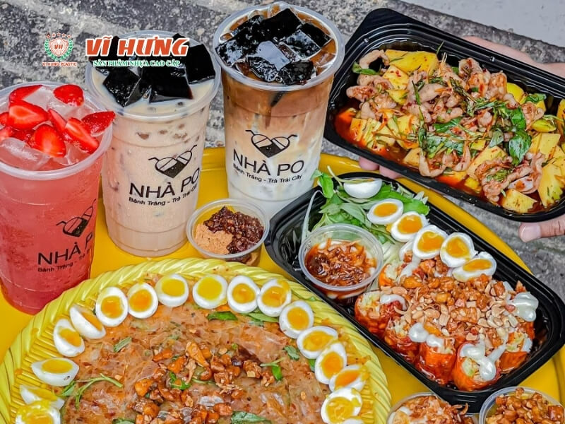 Hai ly trà sữa "Nhà Pô" và các đĩa đồ ăn vặt gồm bánh tráng trộn được trang trí với trứng cút, các loại que nướng như chả cá và xúc xích, tokbokki cay, sắp xếp trên bàn với các chén nước mắm và gia vị