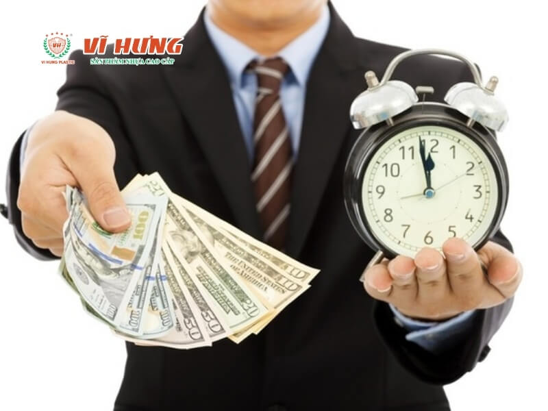 Người đàn ông mặc áo sơ mi xanh và cà vạt nâu cầm tờ tiền đô la Mỹ và đồng hồ báo thức đen, biểu thị khái niệm thời gian và tiền bạc trong kinh doanh