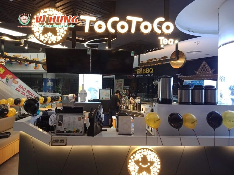 Quán trà sữa ToCoToco tại trung tâm thương mại với biển hiệu neon trắng sáng, quầy phục vụ hiện đại, thiết bị toàn bộ, logo mascot vàng và đen, không gian rộng rãi sạch sẽ
