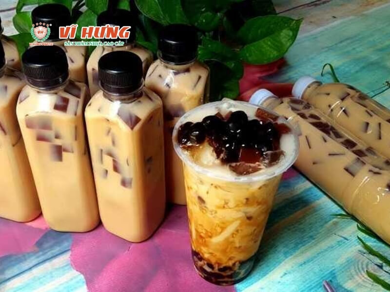 Nhiều chai trà sữa được đựng trong các chai nhựa có nắp đen, một ly trà sữa trân châu đã pha chế sẵn được trưng bày, và các chai chứa trà sữa được chuẩn bị để giao hàng