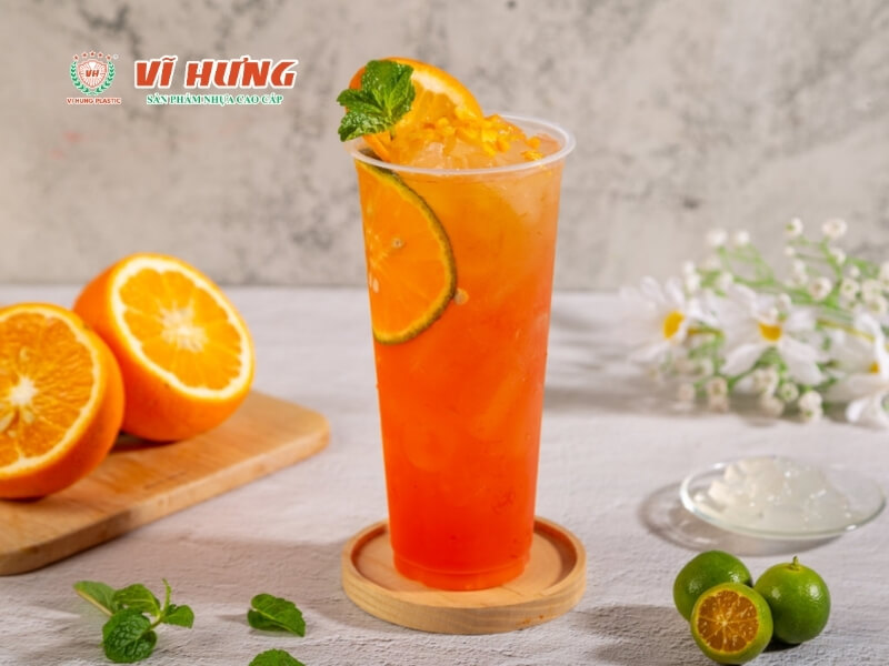 Một ly nước trái cây cam đỏ lạnh với đá, lát cam, lá bạc hà trang trí, đặt trên đĩa gỗ tròn, bên cạnh là cam cắt làm đôi và lime, nền xám đá tự nhiên