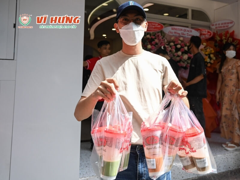 Nhân viên cầm túi giao hàng chứa các ly trà sữa được đóng gói trong túi trong suốt, đứng trước cửa hàng khai trương với nhiều khách hàng phía sau