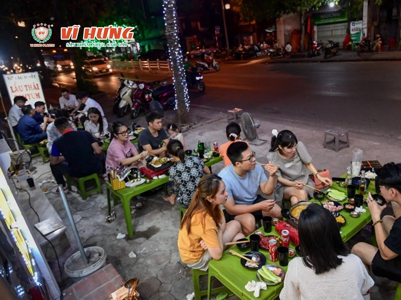 Nhóm bạn trẻ ngồi ăn lẩu nướng vỉa hè trên bàn ghế nhựa xanh dưới ánh đèn đường buổi tối, không khí vui vẻ đặc trưng của quán nhậu bình dân.