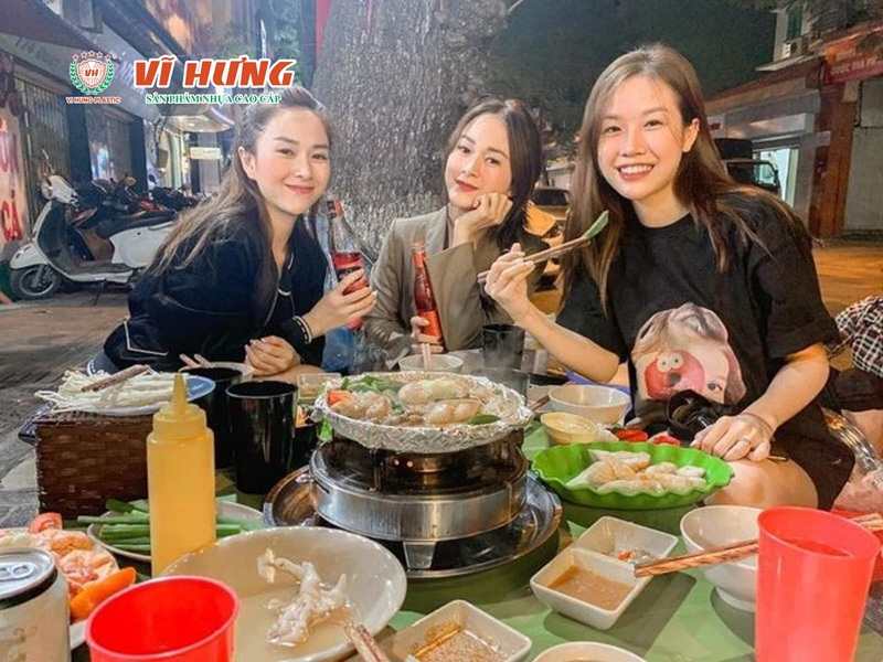 Nhóm bạn nữ đang vui vẻ thưởng thức lẩu tại quán nhỏ với nồi lẩu, rau và các món ăn kèm bày trên bàn.