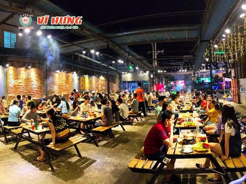 Không gian quán lẩu nướng trong nhà đông khách với hệ thống bàn bếp tích hợp, đèn trang trí và nhiều bàn ăn đang hoạt động trong giờ cao điểm.