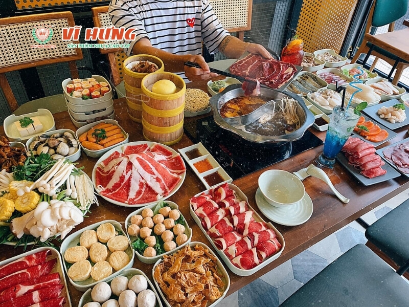 Bàn buffet lẩu nướng với nhiều loại thịt bò, hải sản, viên thả lẩu và rau ăn kèm, thực khách đang gắp thịt nhúng vào nồi lẩu chia ngăn.