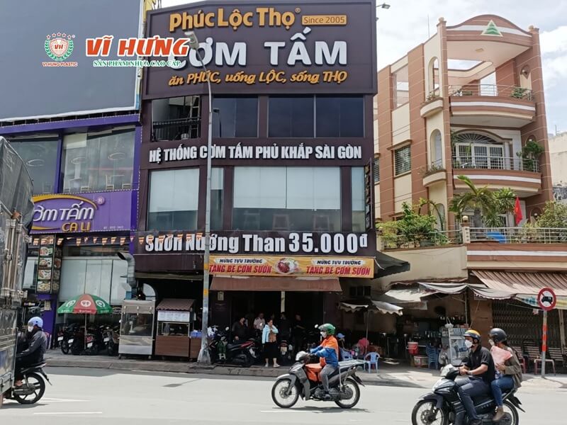 Mặt tiền chi nhánh cơm tấm Phúc Lộc Thọ với biển hiệu lớn nổi bật, bảng giá sườn nướng than và băng rôn khuyến mãi trên con đường đông xe cộ tại Sài Gòn