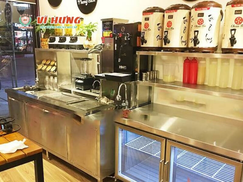 Quầy pha chế trà sữa hiện đại với bề mặt inox sạch sẽ, bình ủ trà màu đen ở trên, tủ mát kính bên phải chứa topping, thiết bị pha chế sắp xếp gọn gàng, dễ vận hành