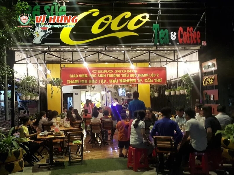 Quán cà phê trà sữa "Coco" tại nông thôn với biển hiệu vàng neon sáng, không gian ngồi ngoài trời rộng rãi, nhiều bàn ghế, khách hàng đông đúc vào buổi tối, cây xanh trang trí quanh quán