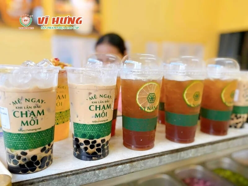 Hàng loạt ly trà sữa được xếp trên quầy với bao bì có logo "Mỗi Ngày Chậm Mỗi", chứa trà nâu và trân châu đen, khách hàng nữ đứng phía sau quầy phục vụ