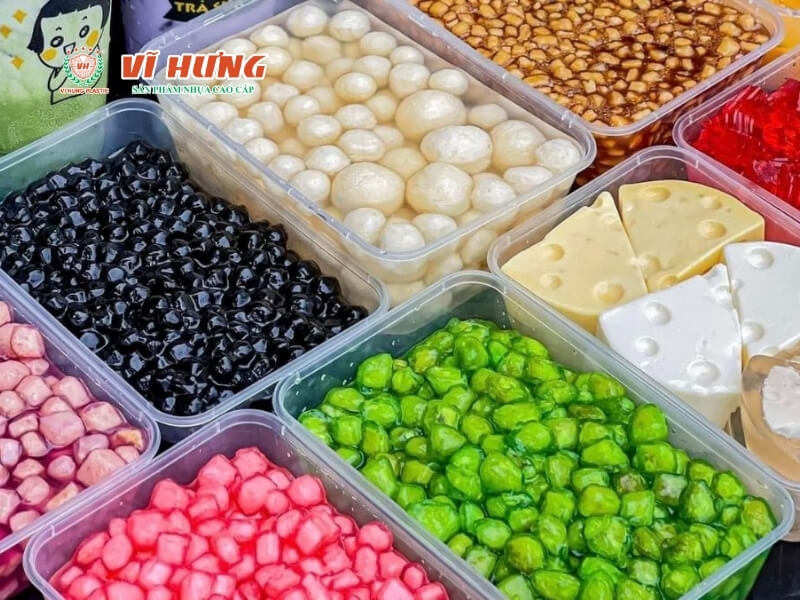 Nhiều hộp nhựa trong suốt chứa các loại topping trà sữa đa sắc gồm trân châu hồng, đen, trắng, thạch xanh lá, đậu đỏ, và các loại topping khác được sắp xếp gọn gàng