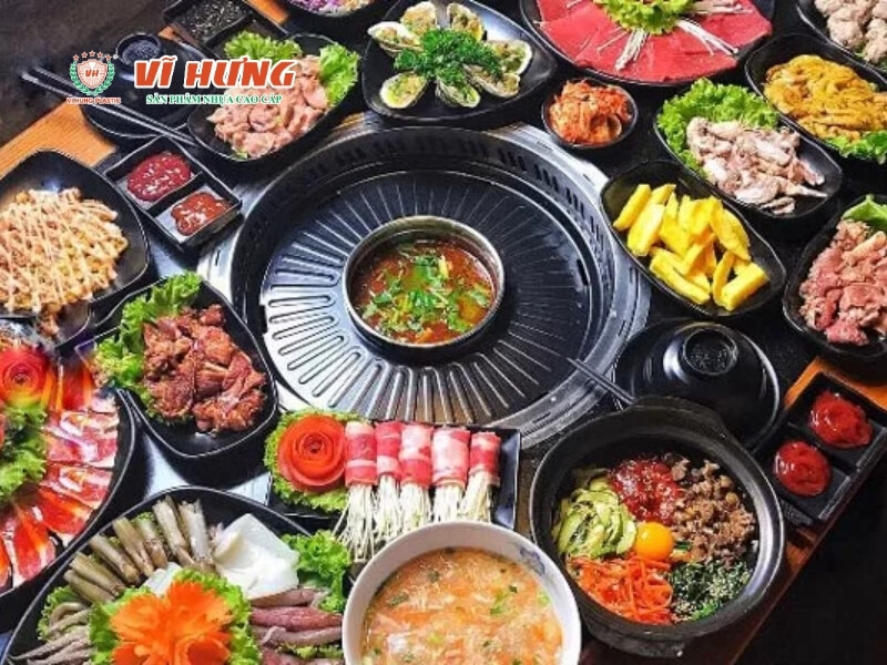 Bàn buffet lẩu nướng không khói với bếp nướng trung tâm, xung quanh là nhiều đĩa thịt, hải sản, rau và món ăn kèm được bày kín.